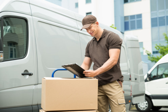 Young Happy Delivery Man With Cardboard Boxes Writing On Clipboard
